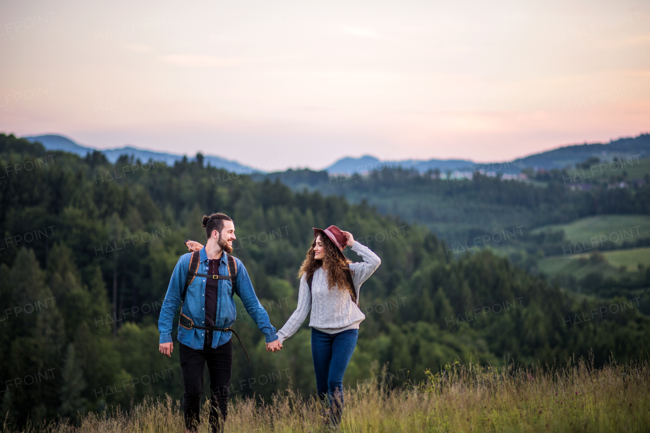 Young tourist couple travellers with backpacks hiking in nature at dusk, holding hands.