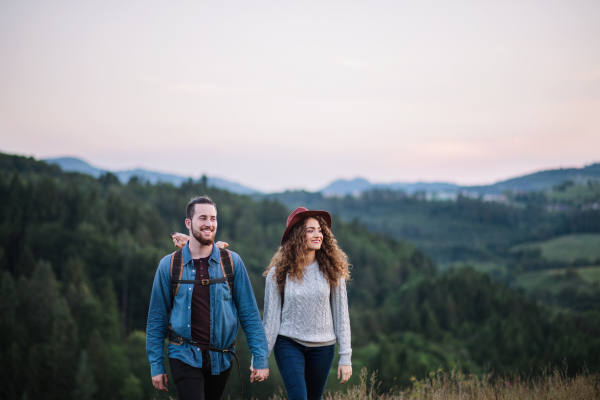 Young tourist couple travellers with backpacks hiking in nature at dusk, holding hands.