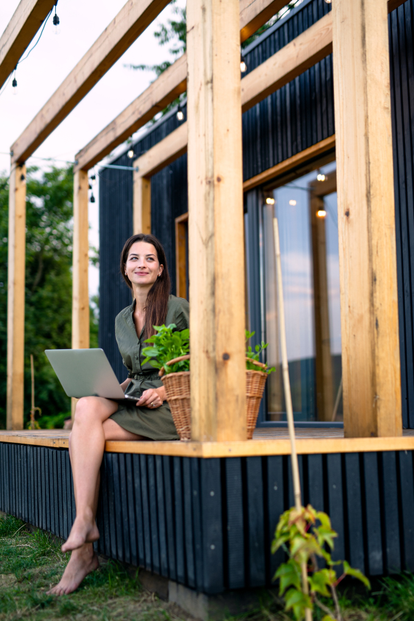 Happy young woman with laptop outdoors, weekend away in container house in countryside.