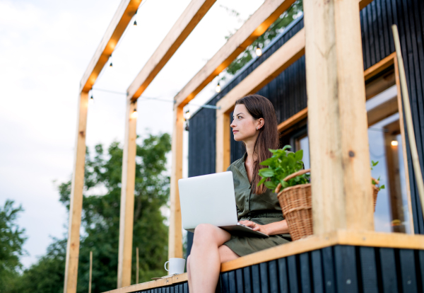 Happy young woman with laptop outdoors, weekend away in container house in countryside.