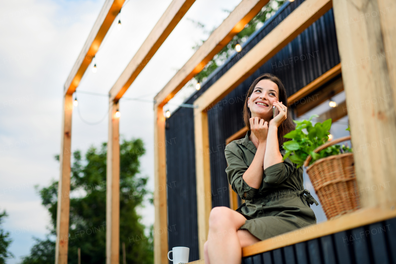 Happy young woman with smartphone outdoors, weekend away in container house in countryside.