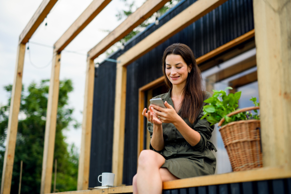 Happy young woman with smartphone outdoors, weekend away in container house in countryside.