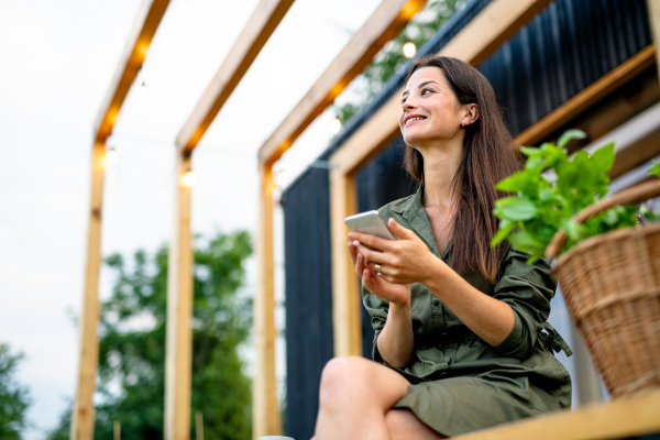 Happy young woman with smartphone outdoors, weekend away in container house in countryside.