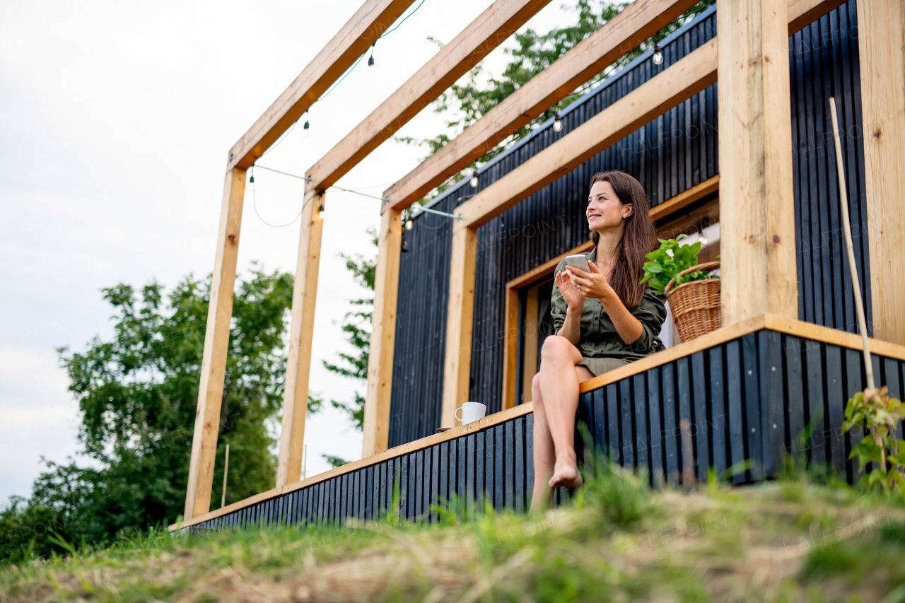 Happy young woman with smartphone outdoors, weekend away in container house in countryside.