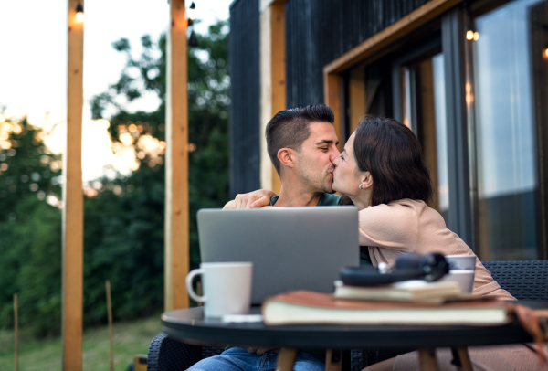 Young couple with laptop sitting and kissing outdoors, weekend away in container house in countryside.