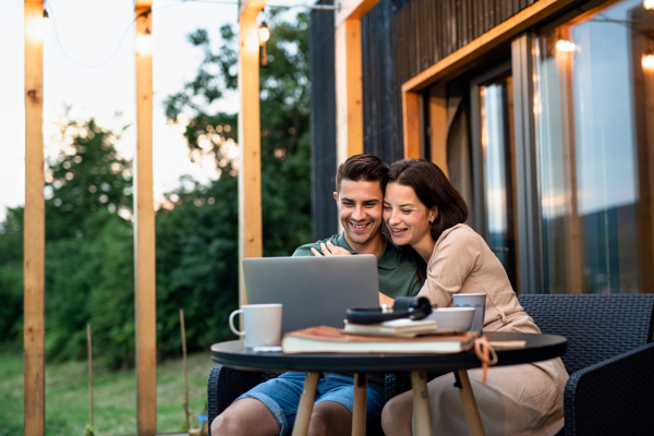 Young couple with laptop sitting outdoors, weekend away in container house in countryside.