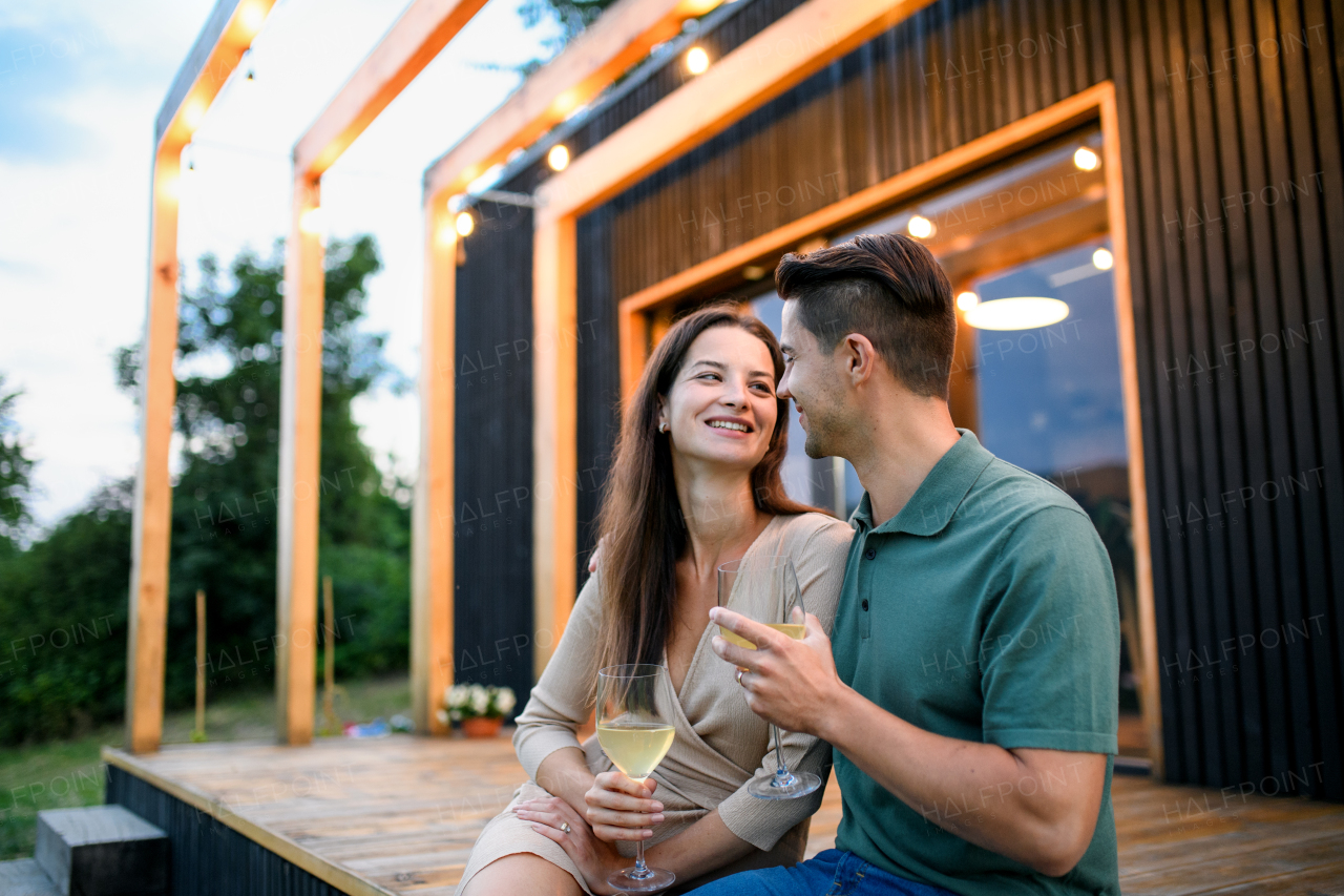 Young couple with wine sitting outdoors, weekend away in container house in countryside.