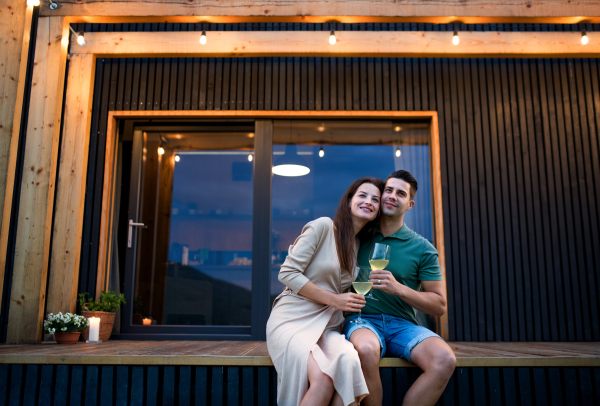 Young couple with wine sitting outdoors, weekend away in container house in countryside.