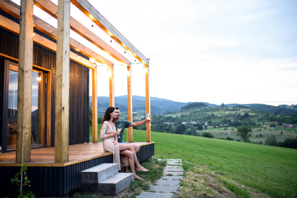 Young couple with wine sitting outdoors, weekend away in container house in countryside.