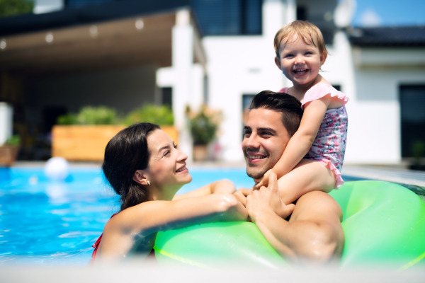 Happy young family with small daughter in swimming pool outdoors in backyard garden.