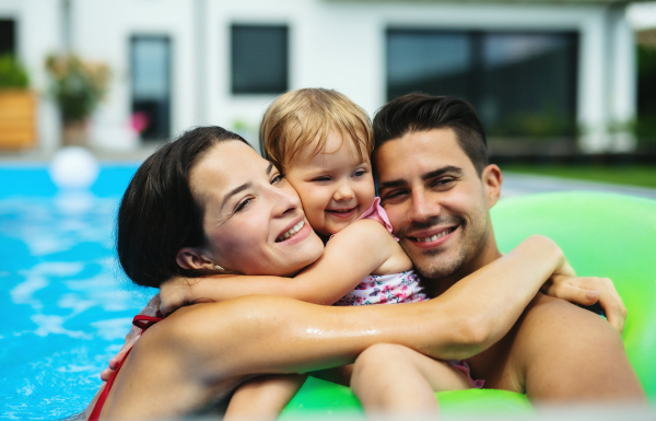 Happy young family with small daughter in swimming pool outdoors in backyard garden.