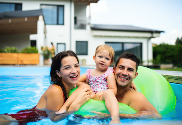 Happy young family with small daughter in swimming pool outdoors in backyard garden.