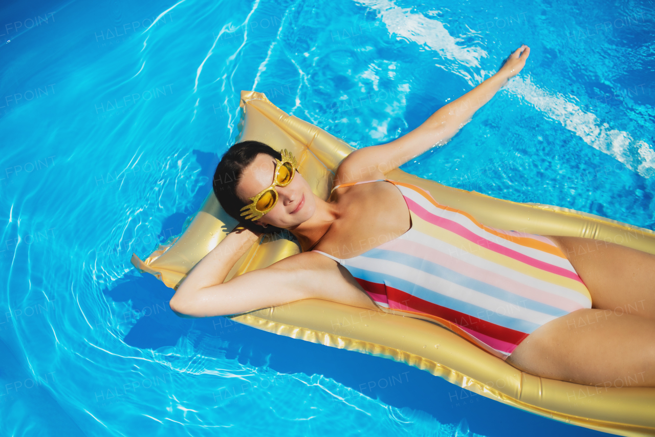 Top view of young woman with sunglasses in swimming pool outdoors on floating bed, relaxing.