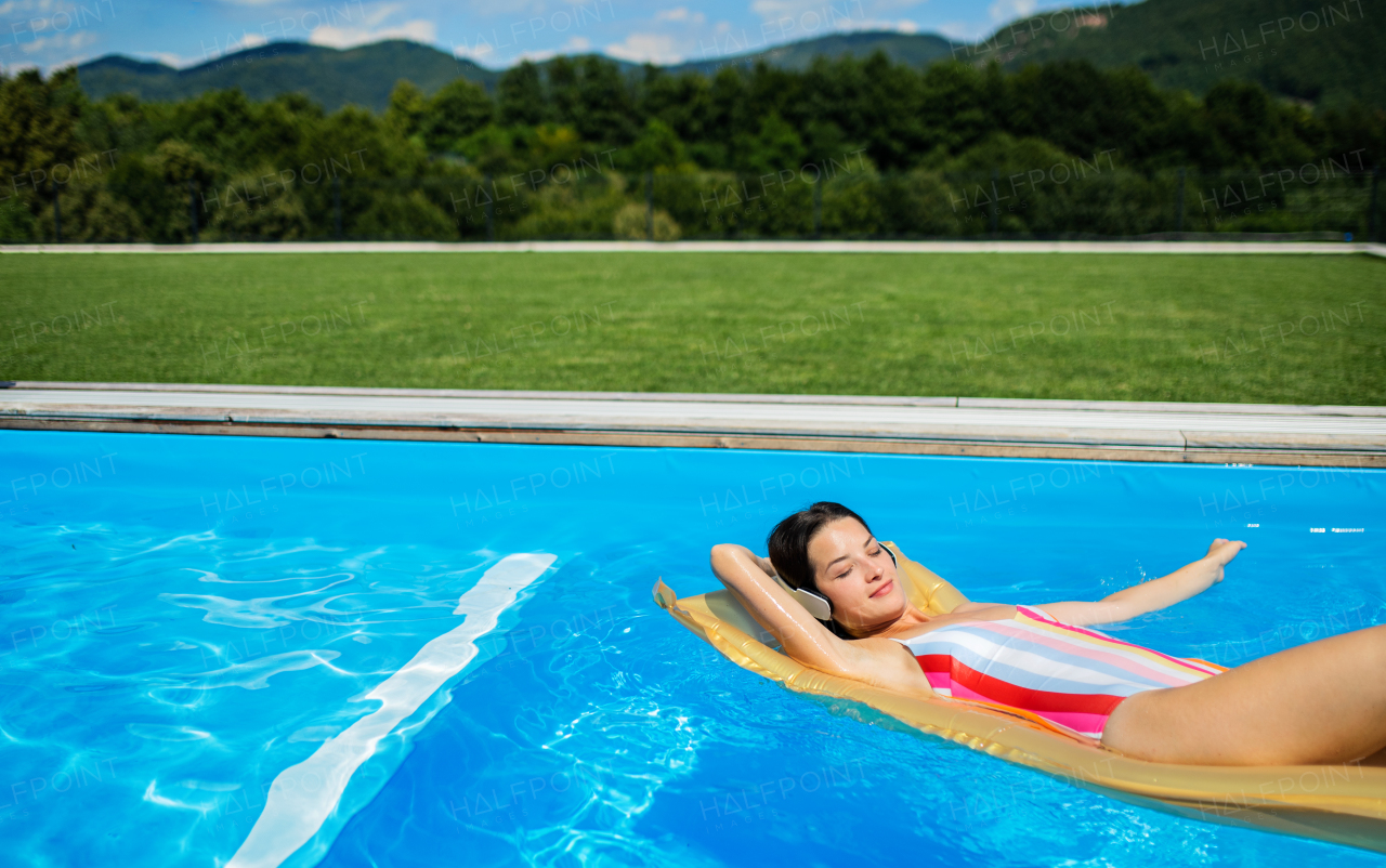 Young woman with headphones in swimming pool outdoors in backyard garden, relaxing.