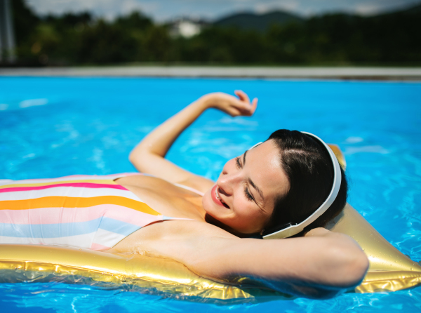 Young woman with headphones in swimming pool outdoors in backyard garden, relaxing.