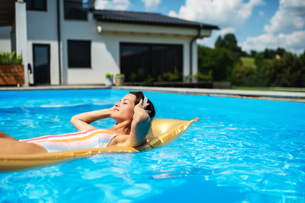 Young woman with headphones in swimming pool outdoors in backyard garden, relaxing.