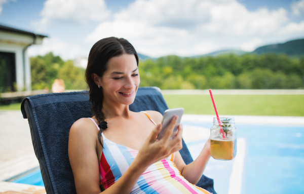 Young woman with drink sitting by swimming pool outdoors in backyard garden, relaxing.