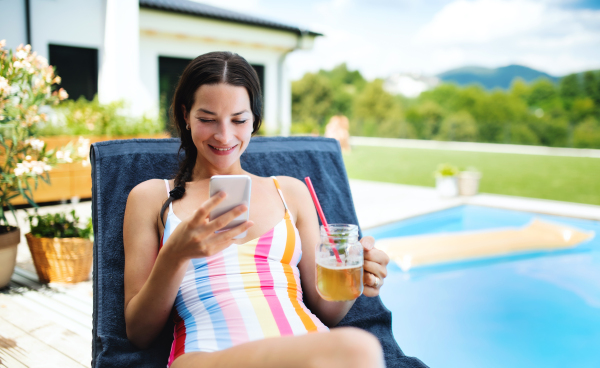 Beautiful young woman sitting by swimming pool outdoors in backyard garden, using smartphone.