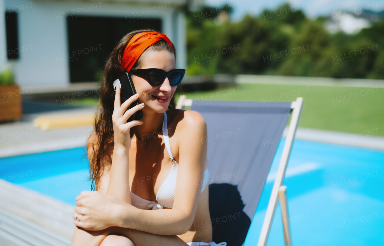 A young woman sitting by swimming pool outdoors in backyard garden, making a call.