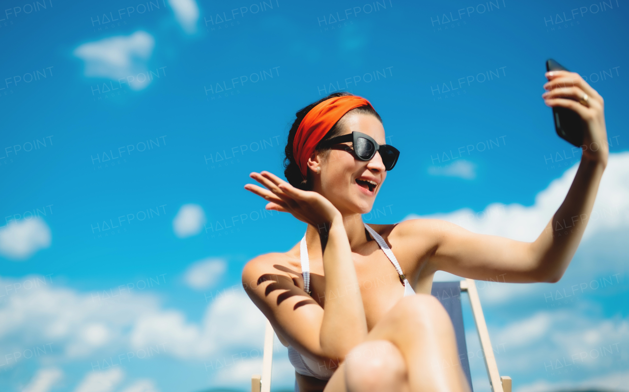 Young woman sitting by swimming pool outdoors in backyard garden, taking selfie with smartphone.