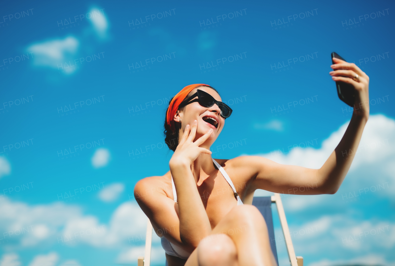 Young woman sitting by swimming pool outdoors in backyard garden, taking selfie with smartphone.