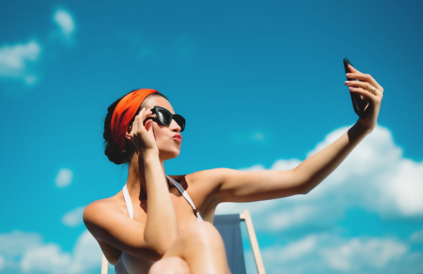 Young woman sitting by swimming pool outdoors in backyard garden, taking selfie with smartphone.