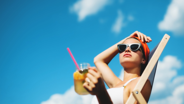 Young woman with drink sitting by swimming pool outdoors in backyard garden, relaxing.