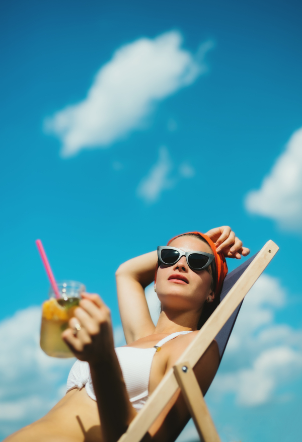 Young woman with drink sitting by swimming pool outdoors in backyard garden, relaxing.