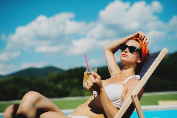 Young woman with drink sitting by swimming pool outdoors in backyard garden, relaxing.