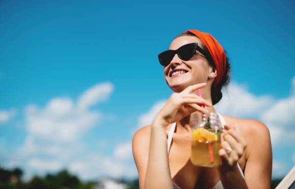 Young woman with drink sitting by swimming pool outdoors in backyard garden, relaxing.