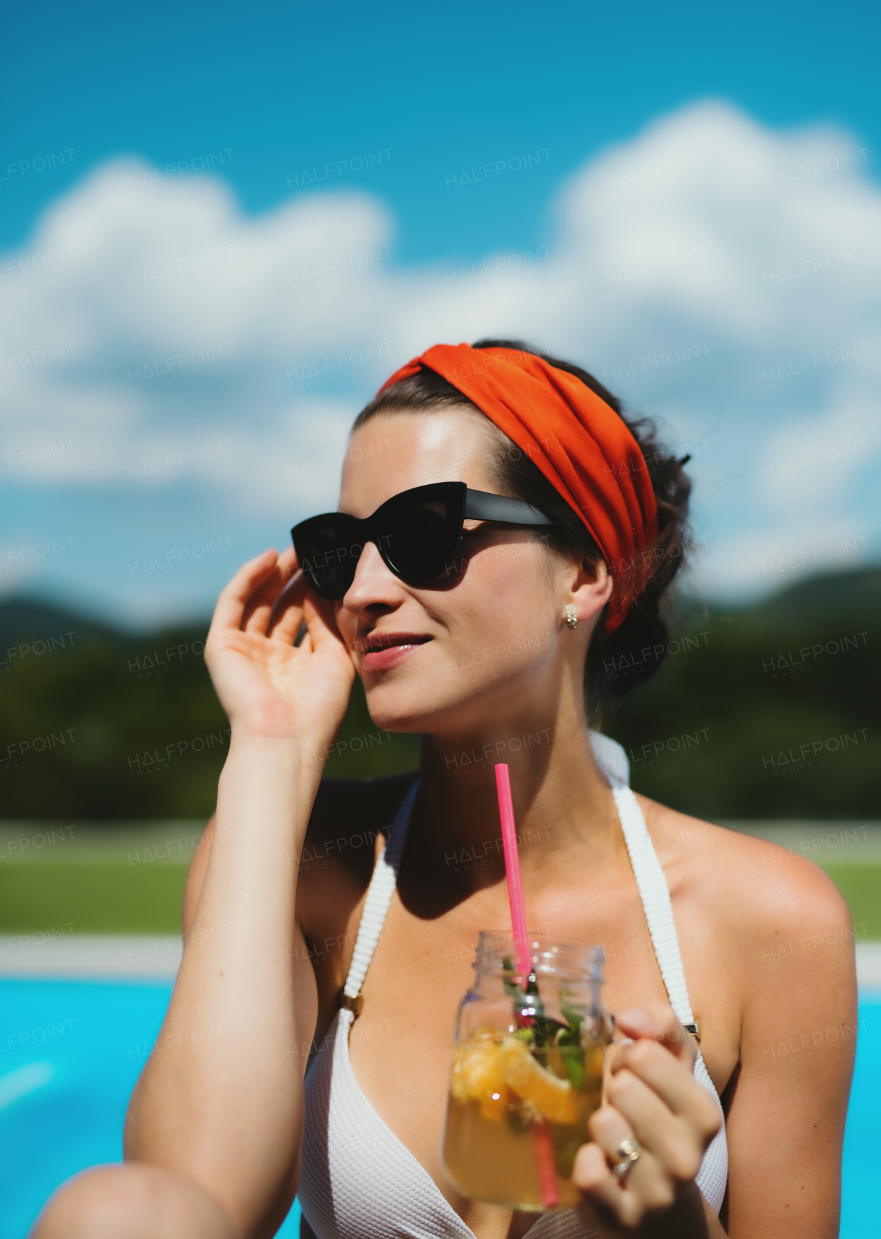 Young woman with drink sitting by swimming pool outdoors in backyard garden, relaxing.