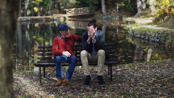 Senior father and his young son sitting on bench by lake in nature, talking.