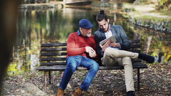 A senior father and his son sitting on bench in nature, using tablet.