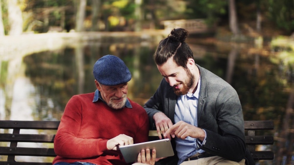 A senior father and his son sitting on bench in nature, using tablet.