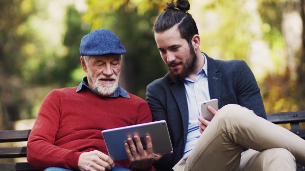 A senior father and his son sitting on bench in nature, using tablet and smartphone.