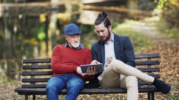 A senior father and his son sitting on bench in nature, using tablet and smartphone.