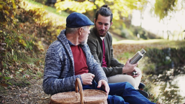 Senior father and his young son sitting by lake in nature, talking and eating.