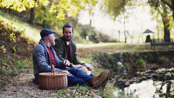 Senior father and his young son sitting by lake in nature, talking and eating.