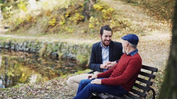 Senior father and his young son sitting on bench by lake in nature, talking.