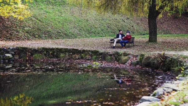 Senior father and his young son sitting on bench by lake in nature, talking.