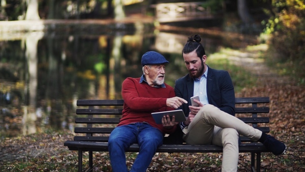 A senior father and his son sitting on bench in nature, using tablet and smartphone. Slow motion.