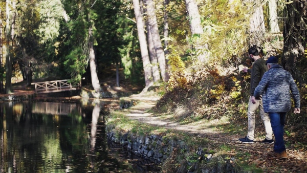 Rearview of senior father and his young son walking by lake in nature, talking. Slow motion.