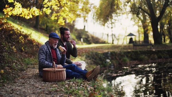 Senior father and his young son sitting by lake in nature, talking and eating. Slow motion.