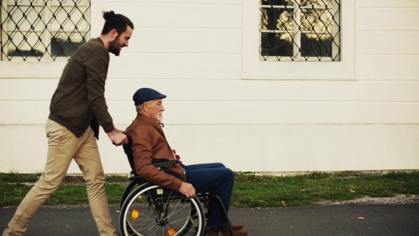 Young man and his senior father in wheelchair on a walk in town, having fun. Slow motion.