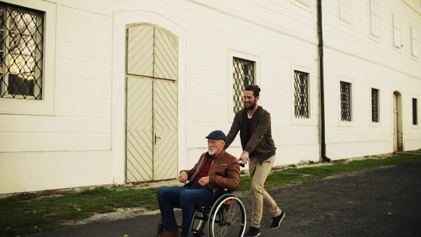 Young man and his senior father in wheelchair on a walk in town, having fun. Slow motion.