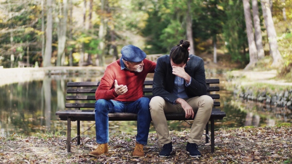 Senior father comforting his sad and frustrated adult son on bench by lake in nature.