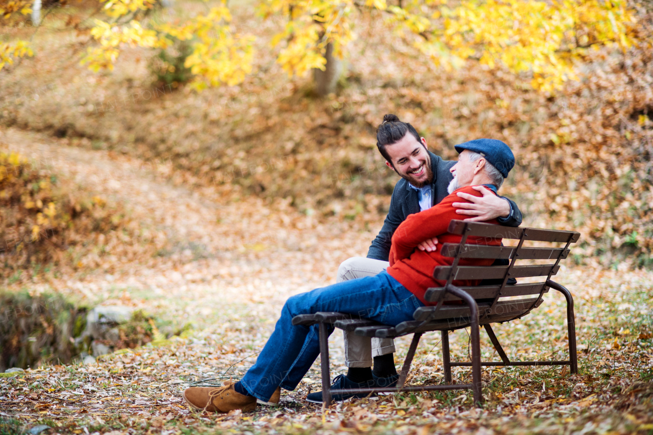 Senior father and his young son sitting on bench by lake in nature, talking.