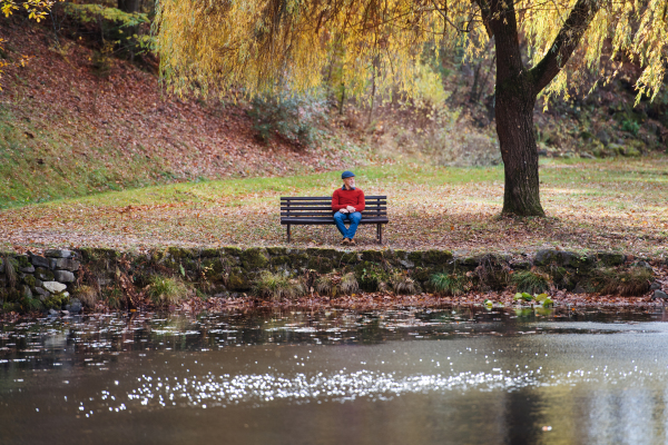 A lonely senior man sitting on bench by lake in nature, looking at camera.