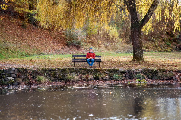 A lonely senior man sitting on bench by lake in nature, looking at camera.