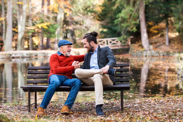 Senior father and his young son sitting on bench by lake in nature, talking.